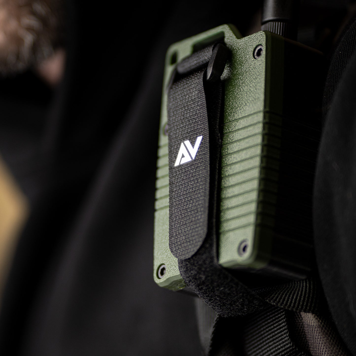 A close-up of a green Atlavox M1 radio, secured to backpack strap on chest.